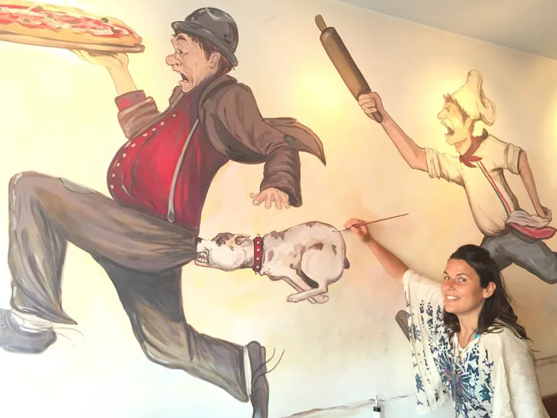 Mural Artist Alessandra Tortone and one of her wall murals of a dog and a chef chasing a man steeling a pizza.